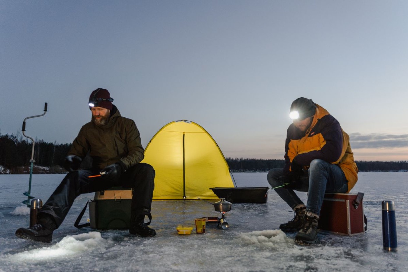 Two anglers fishing beside a glowing shelter at dusk