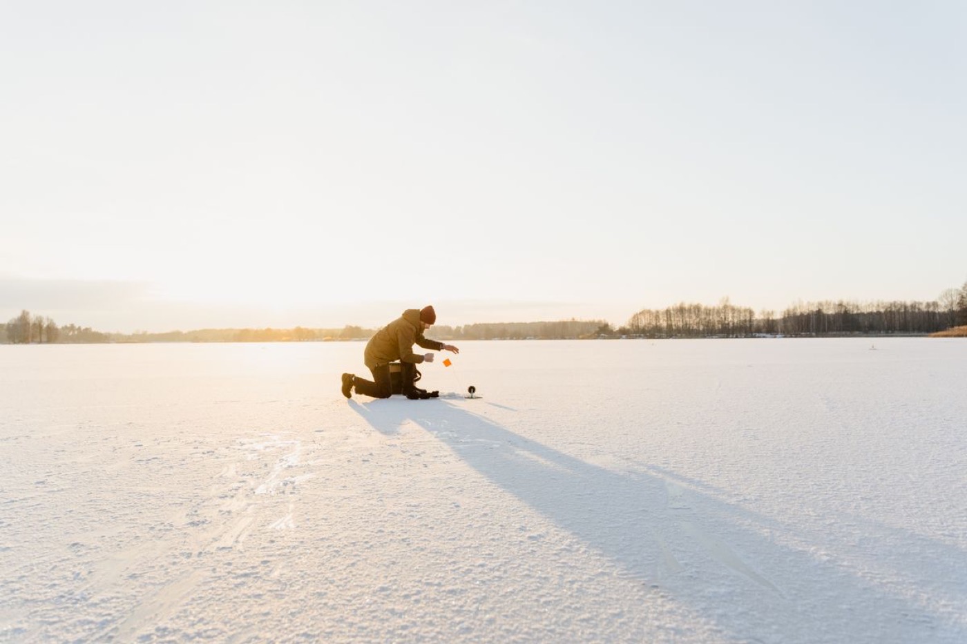 A lone angler kneeling at a hole on a bright frozen lake at sunrise